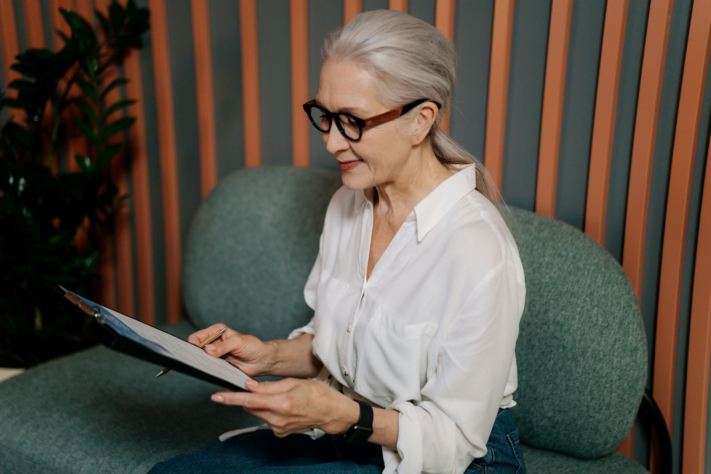 Senior woman sits in her office in white dress shirt, black pants, and eyeglasses looking at clipboard of her employer health insurance information.